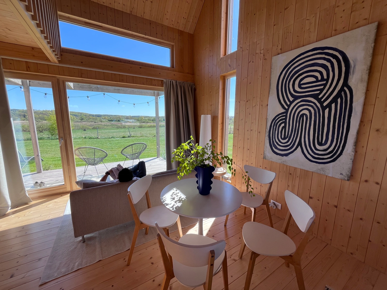 Drewniane wnętrze z dużym oknem na zieloną łąkę, okrągły stół z białymi krzesłami i niebieską wazą z rośliną