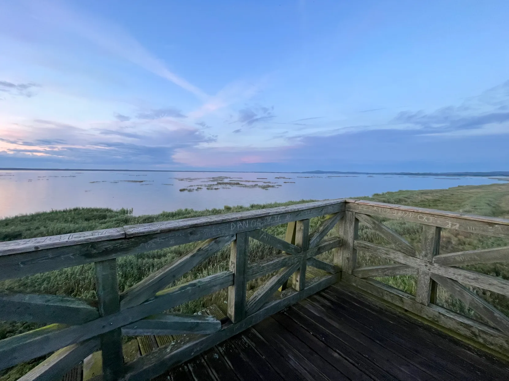 Drewniana platforma z balustradą nad rozległymi trzcinami i spokojnym jeziorem przy pastelowym zmierzchu