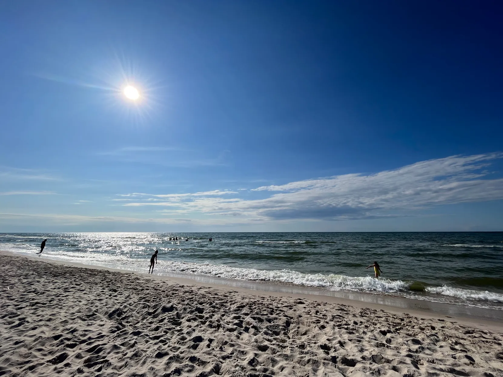 Piaszczysta plaża, błękitne niebo z jasnym słońcem, lśniące morze i kilka osób przy brzegu